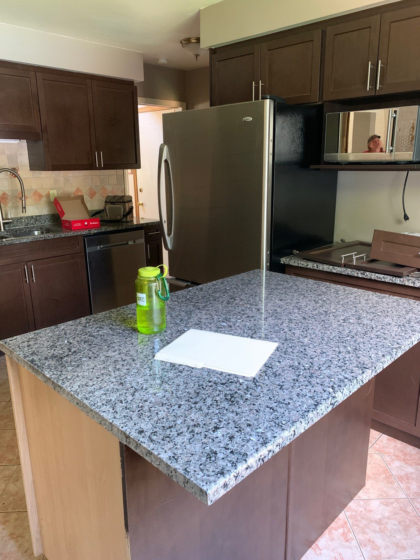 Kitchen with dark wood cabinets, stainless fridge, speckled island, and a green can on the counter.