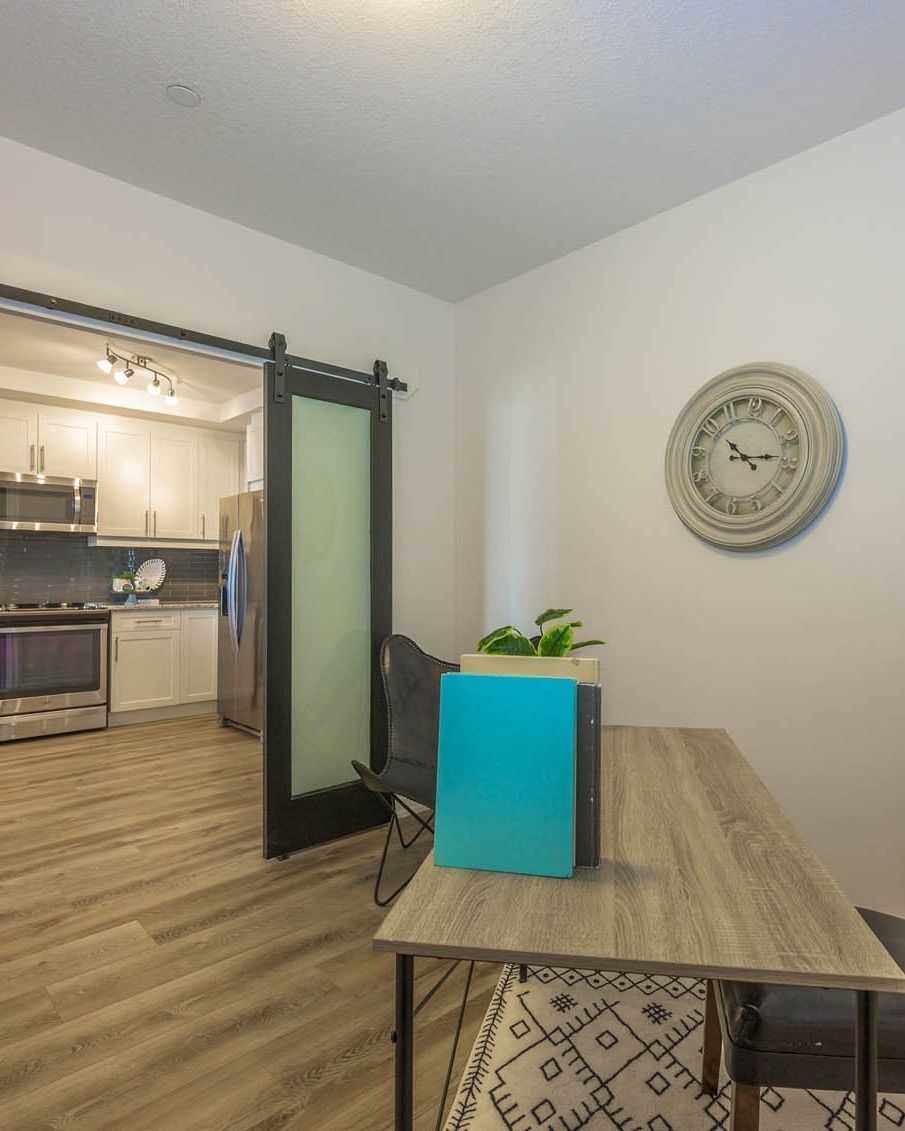 Dining room with wood table, blue notebook, wall clock, and open view into a kitchen.