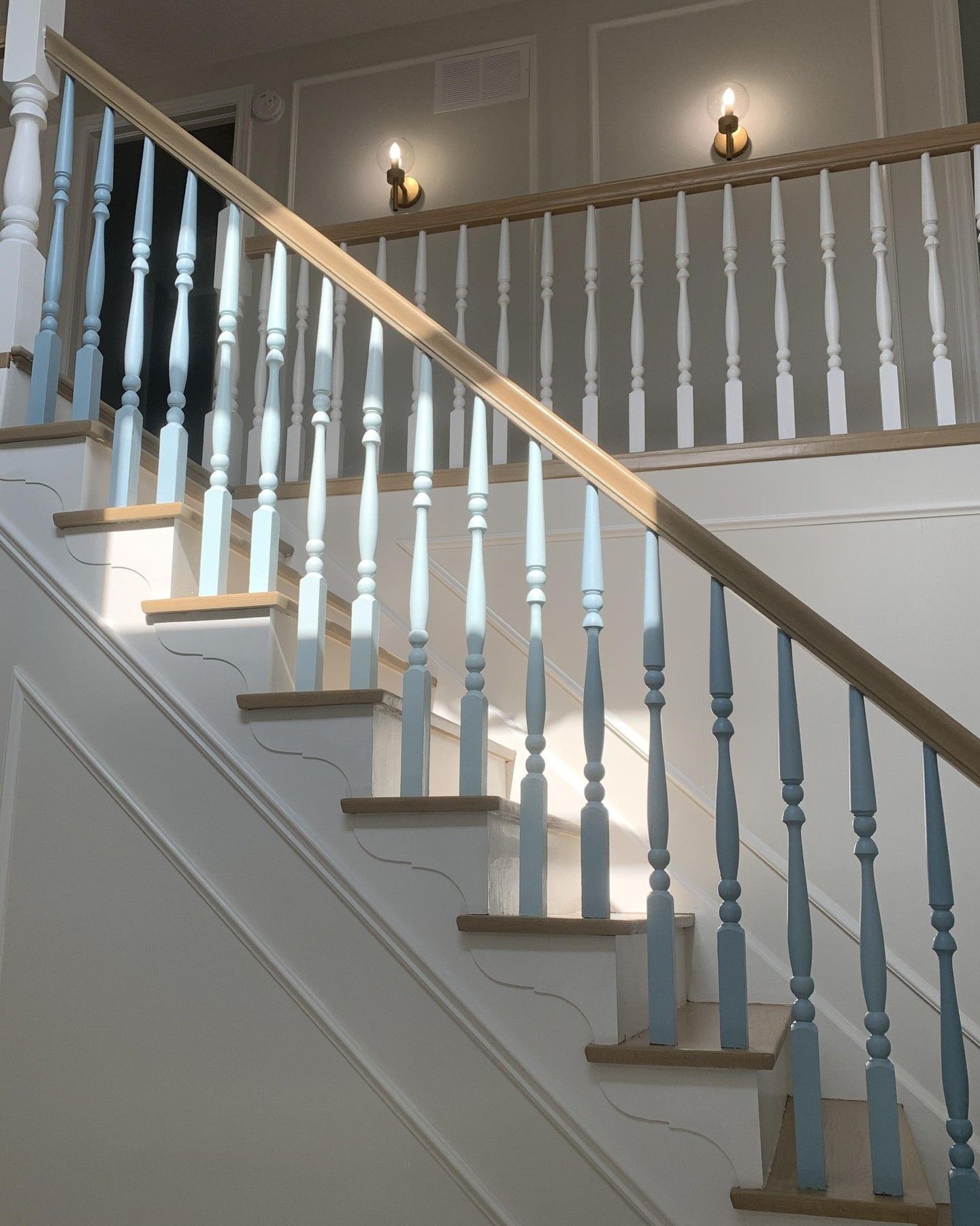 Indoor staircase with white balusters, wooden handrail, and wall sconces on a light gray wall