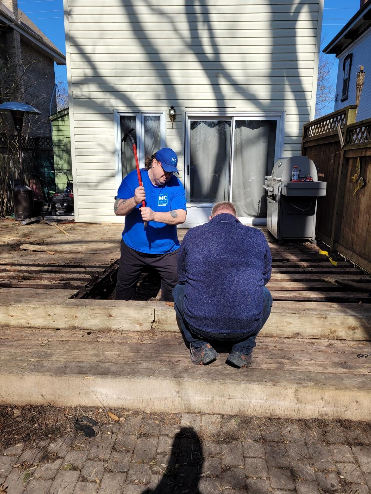 Two people outdoors by a house, one kneeling in a blue shirt and cap, another crouching near the steps.