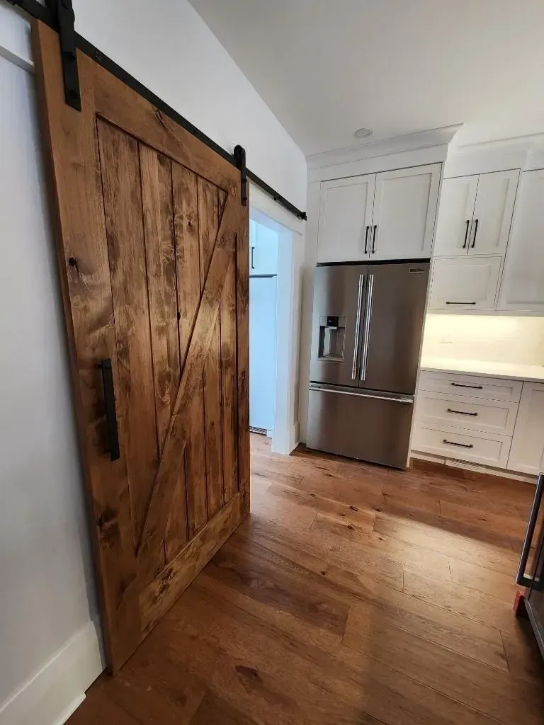 Wooden sliding barn door leading into a bright kitchen with white cabinets and stainless steel fridge.