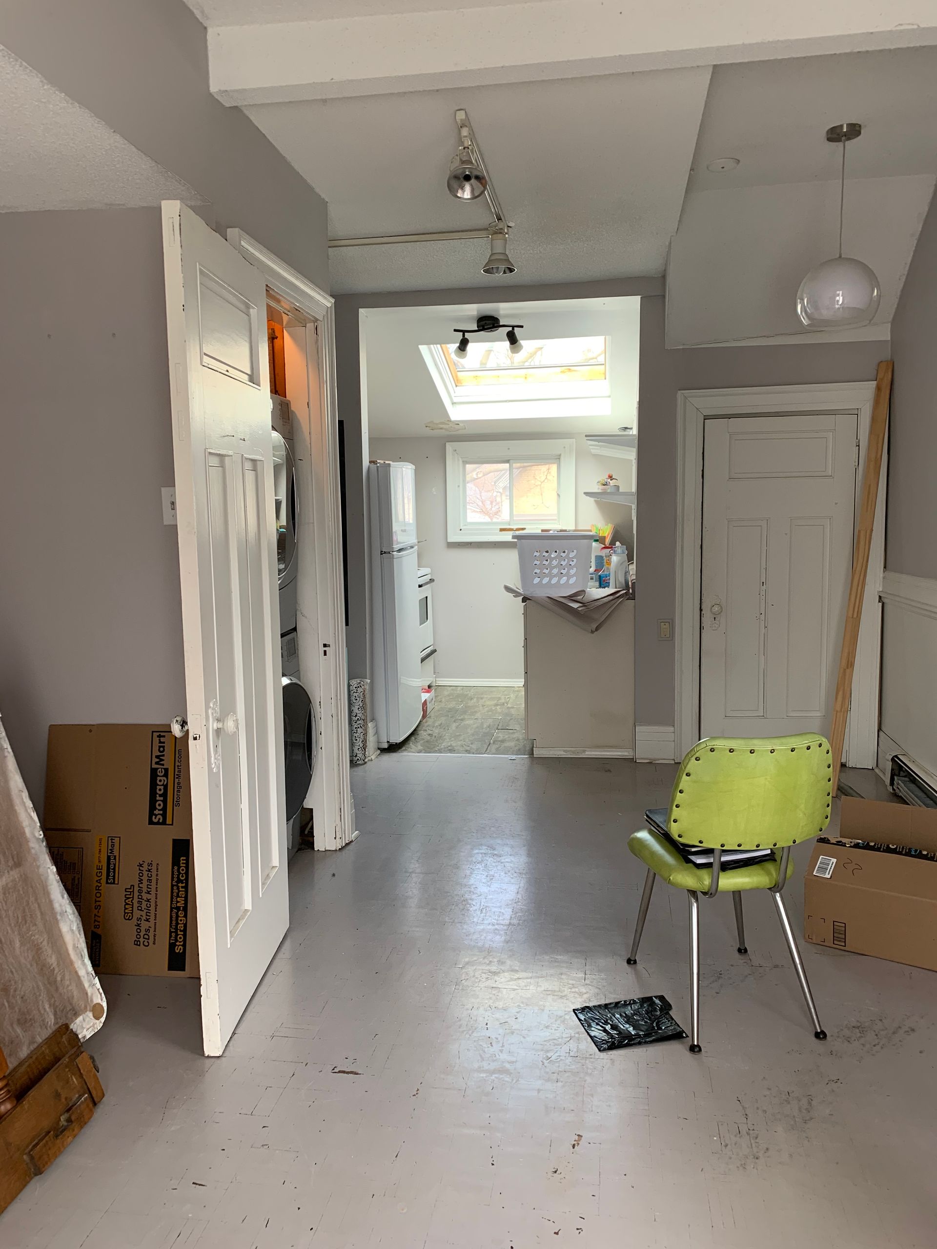 Narrow hallway with white walls, a yellow chair, and a view into a bright kitchen at the end.