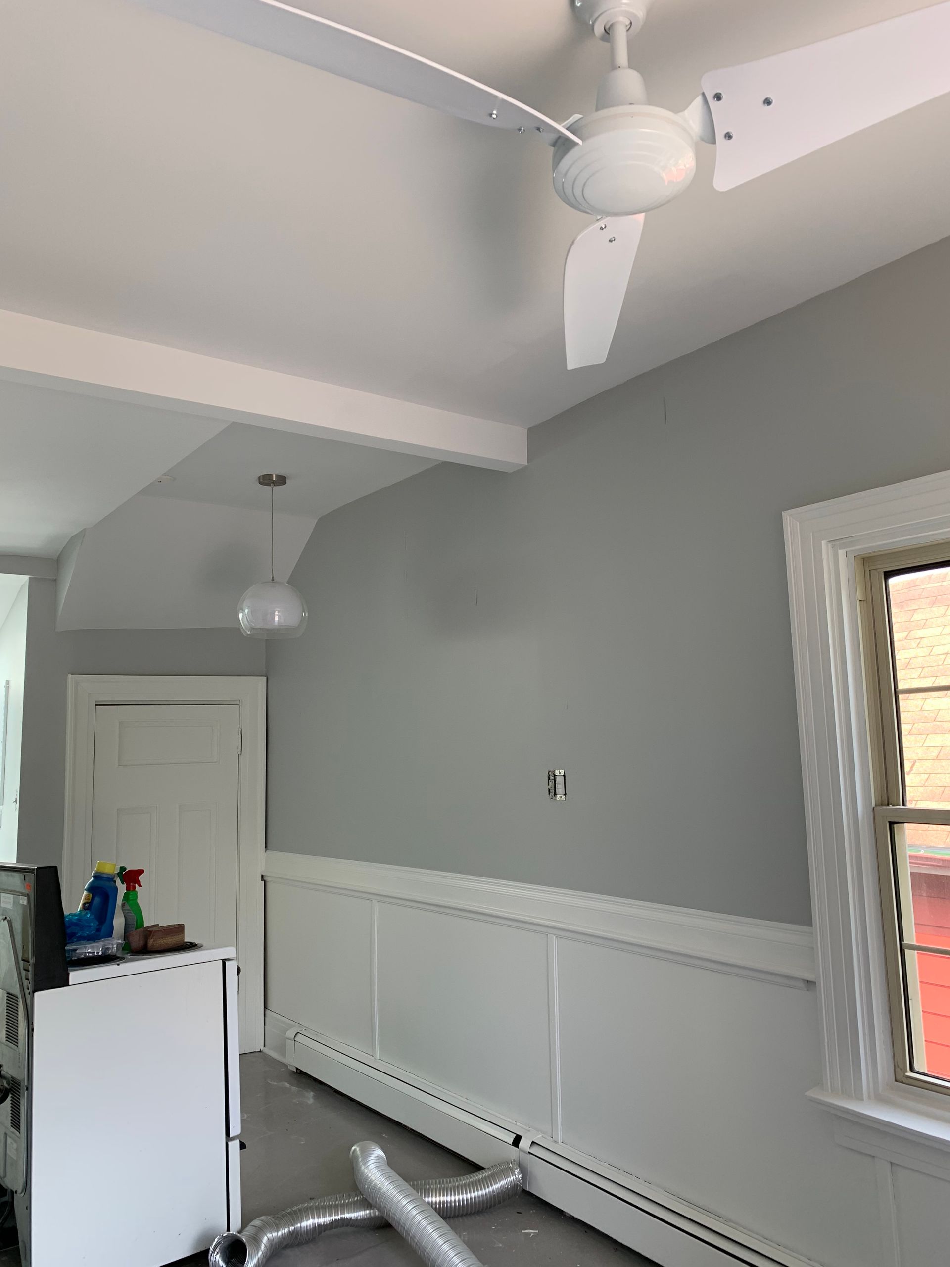 Empty room with gray walls, white wainscoting, ceiling fan, and a window letting in daylight.