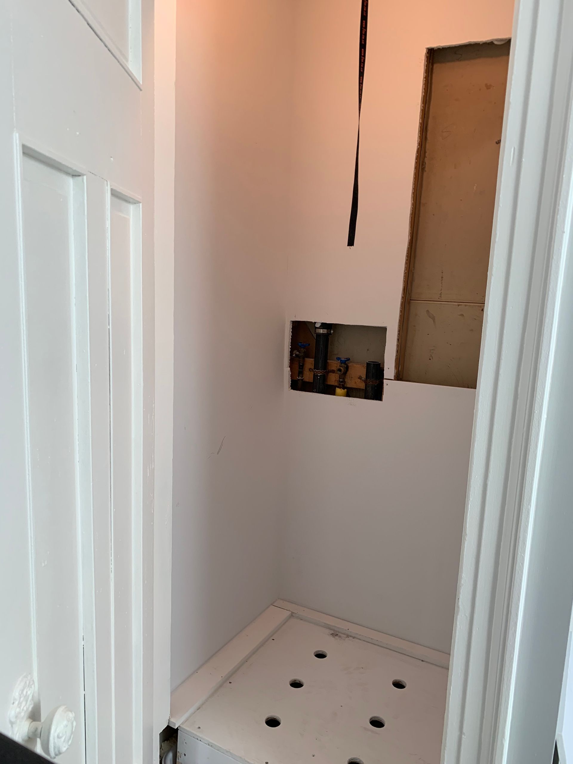 Narrow unfinished shower alcove with white walls, exposed plumbing panel, and tiled floor with drain holes