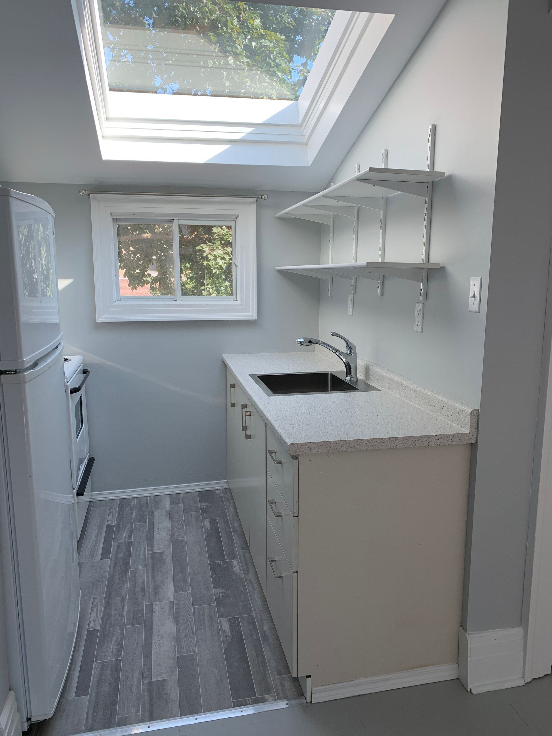 Bright narrow kitchen with skylights, gray flooring, white cabinets, sink, window, and open wall shelves
