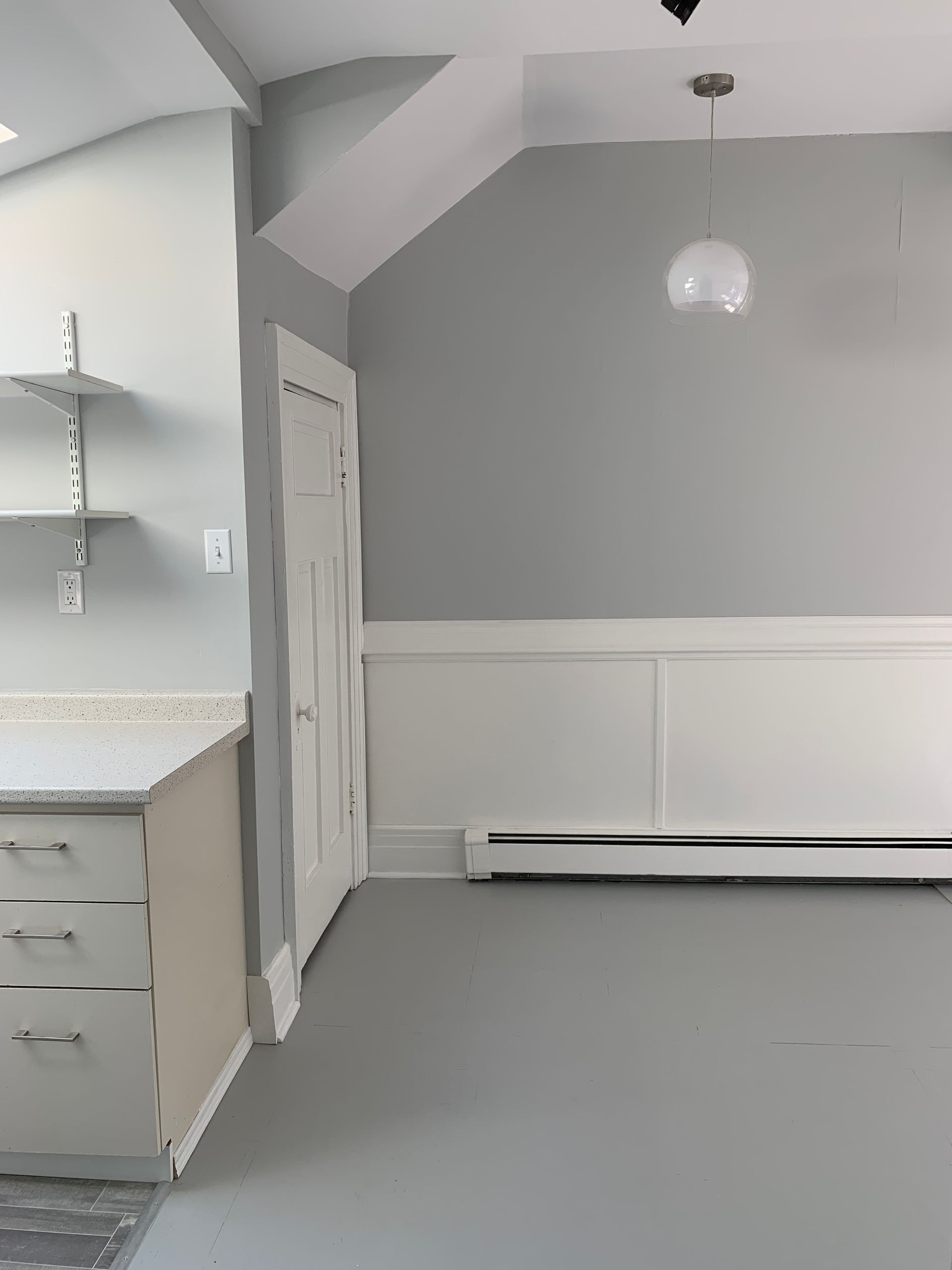 Empty light-gray room with white trim, pendant light, and partial kitchen counter on the left.