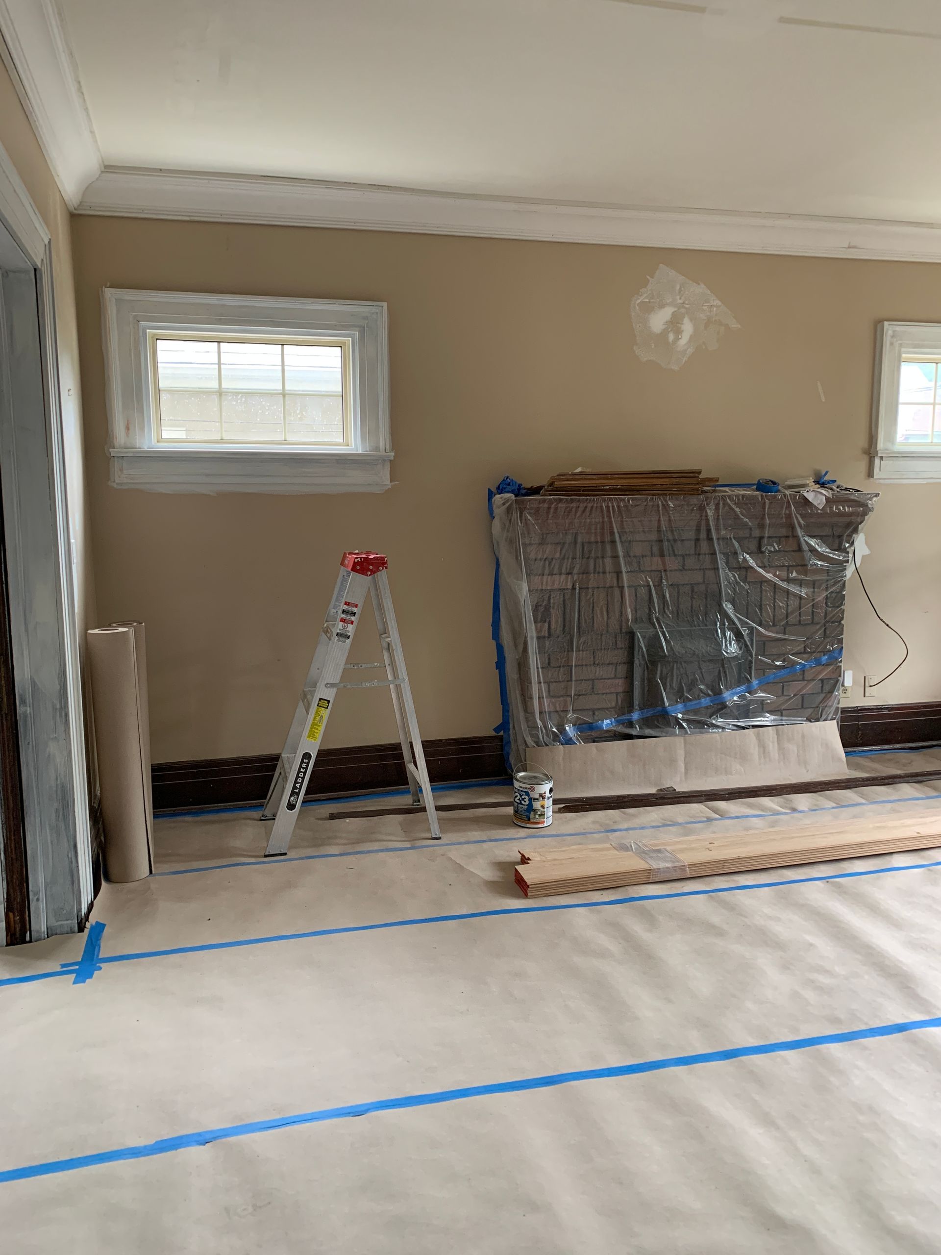 Renovation room with taped floor, ladder, and plastic-covered furniture against beige walls and windows