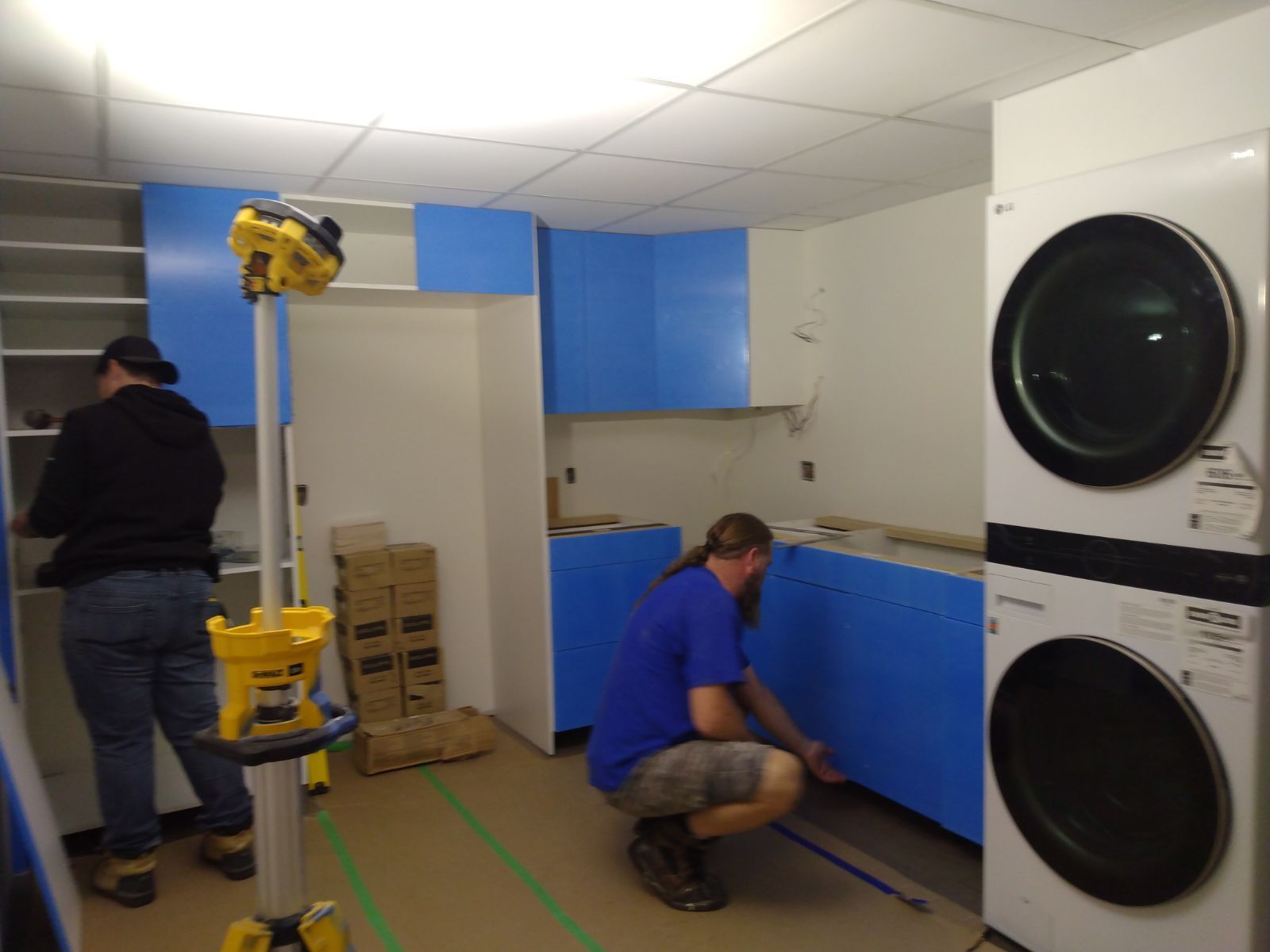 Two workers installing blue cabinets in a room, with stacked washers on the right.