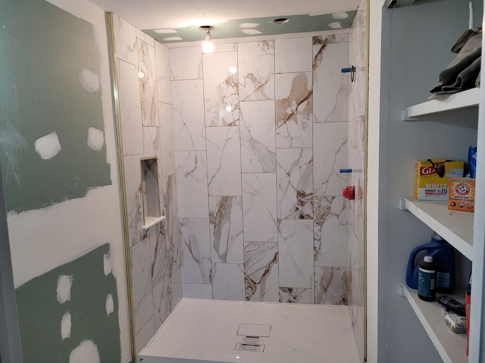 Bathroom under renovation with marble-look tiled shower wall, unfinished drywall, and open shelving