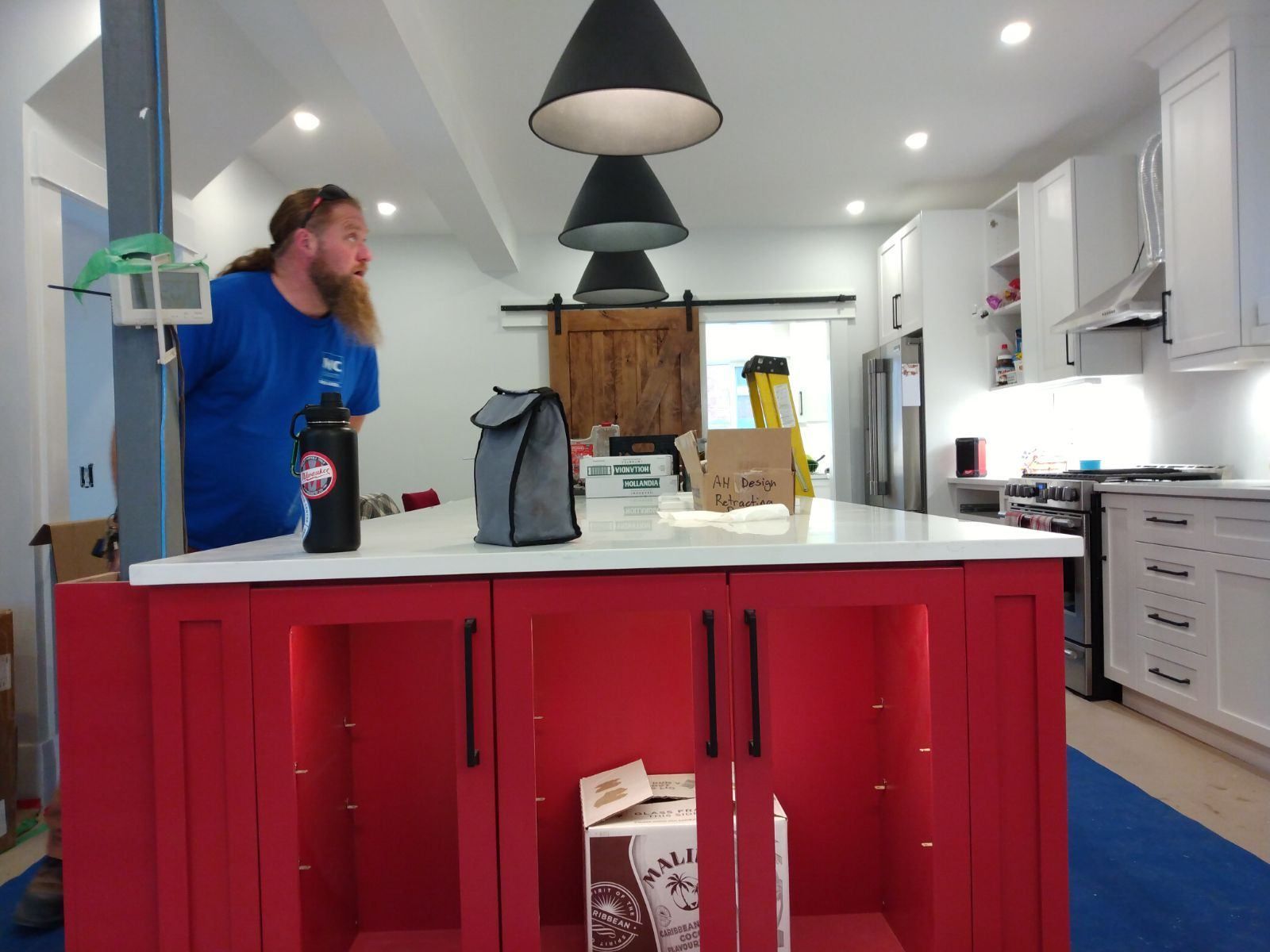 Man in blue shirt in a bright kitchen, leaning over a white island with red cabinets and black pendant lights.