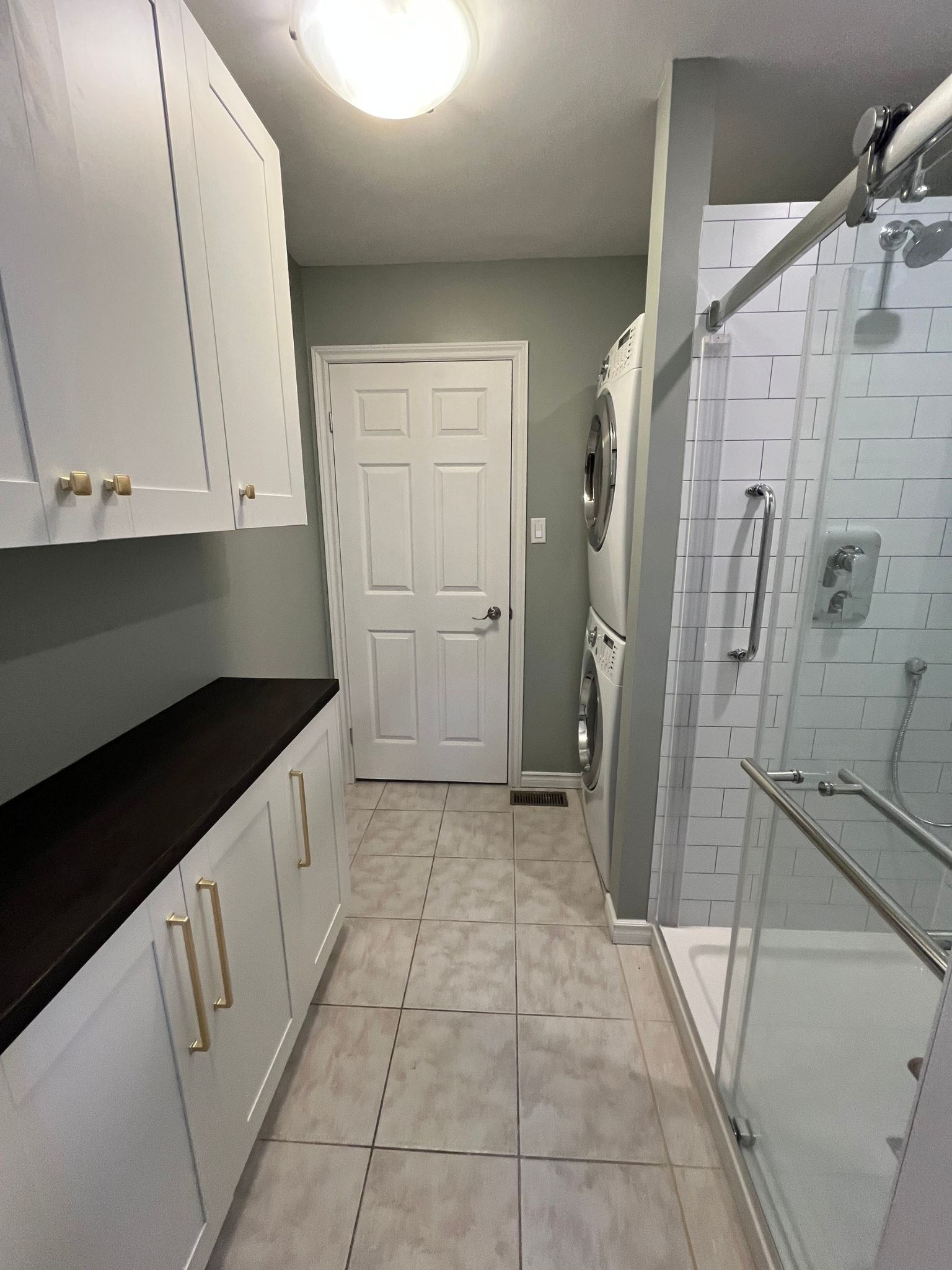 Narrow bathroom with white cabinets, black countertop, tiled floor, and glass shower door.