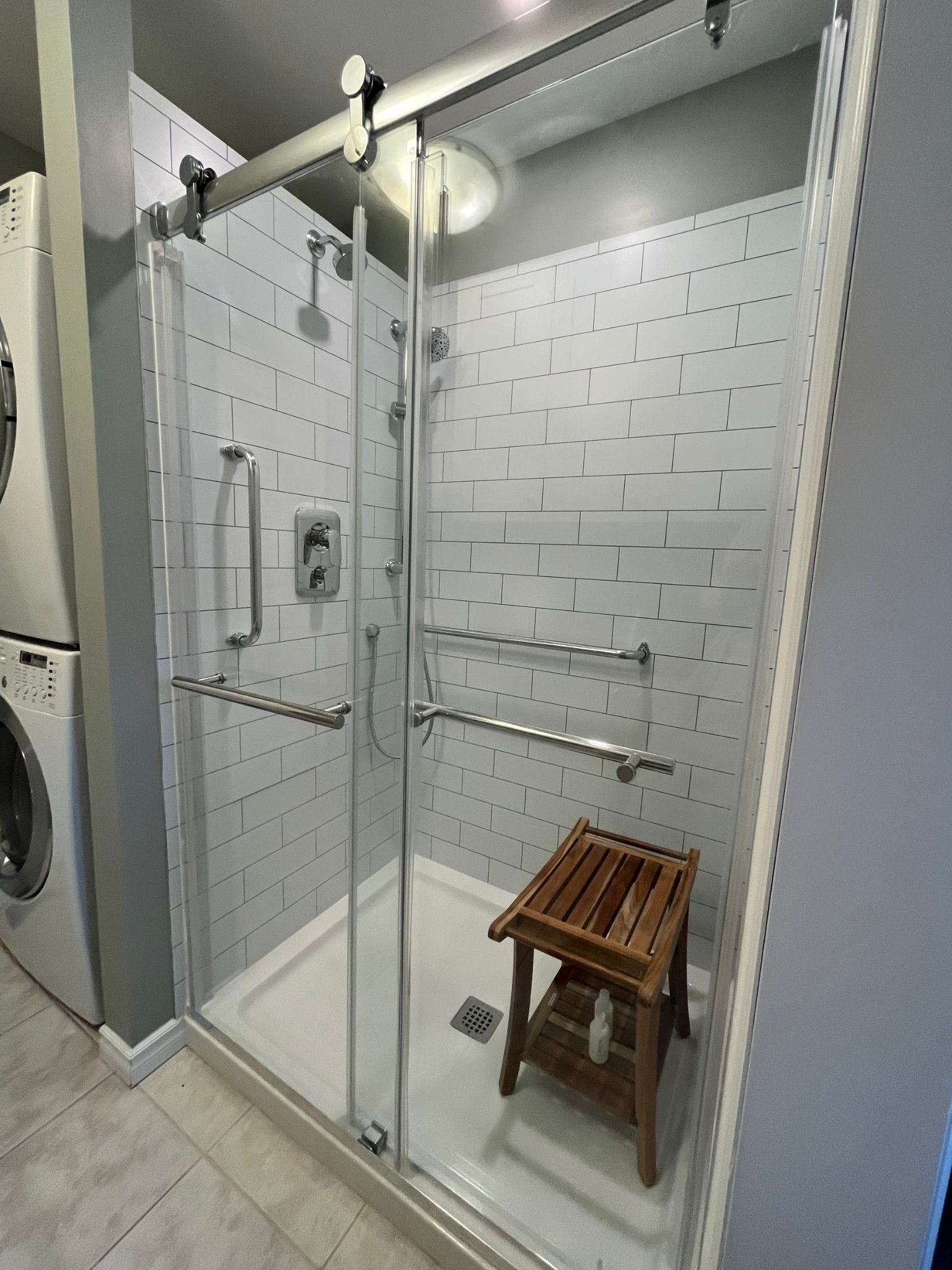 Glass shower stall with chrome grab bars and a wooden shower stool in a white tiled bathroom.
