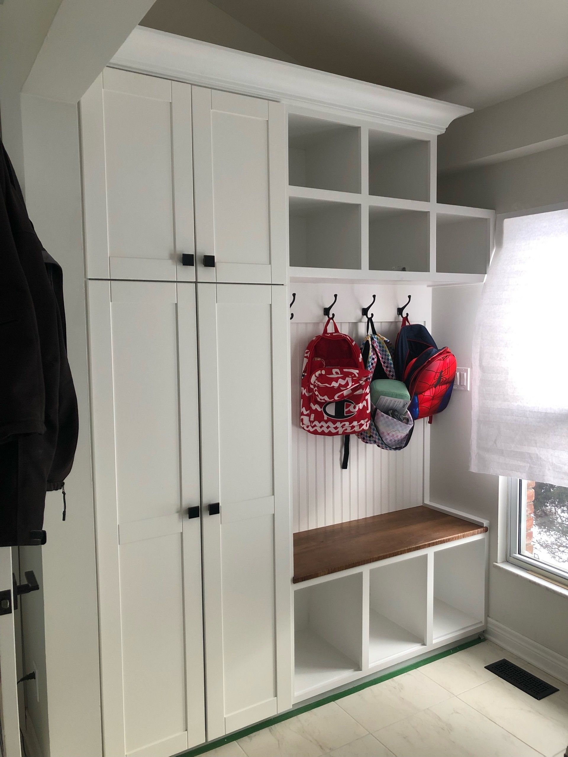 White built-in mudroom cabinet with cubbies, hooks holding backpacks, bench, and closed storage doors by a window.