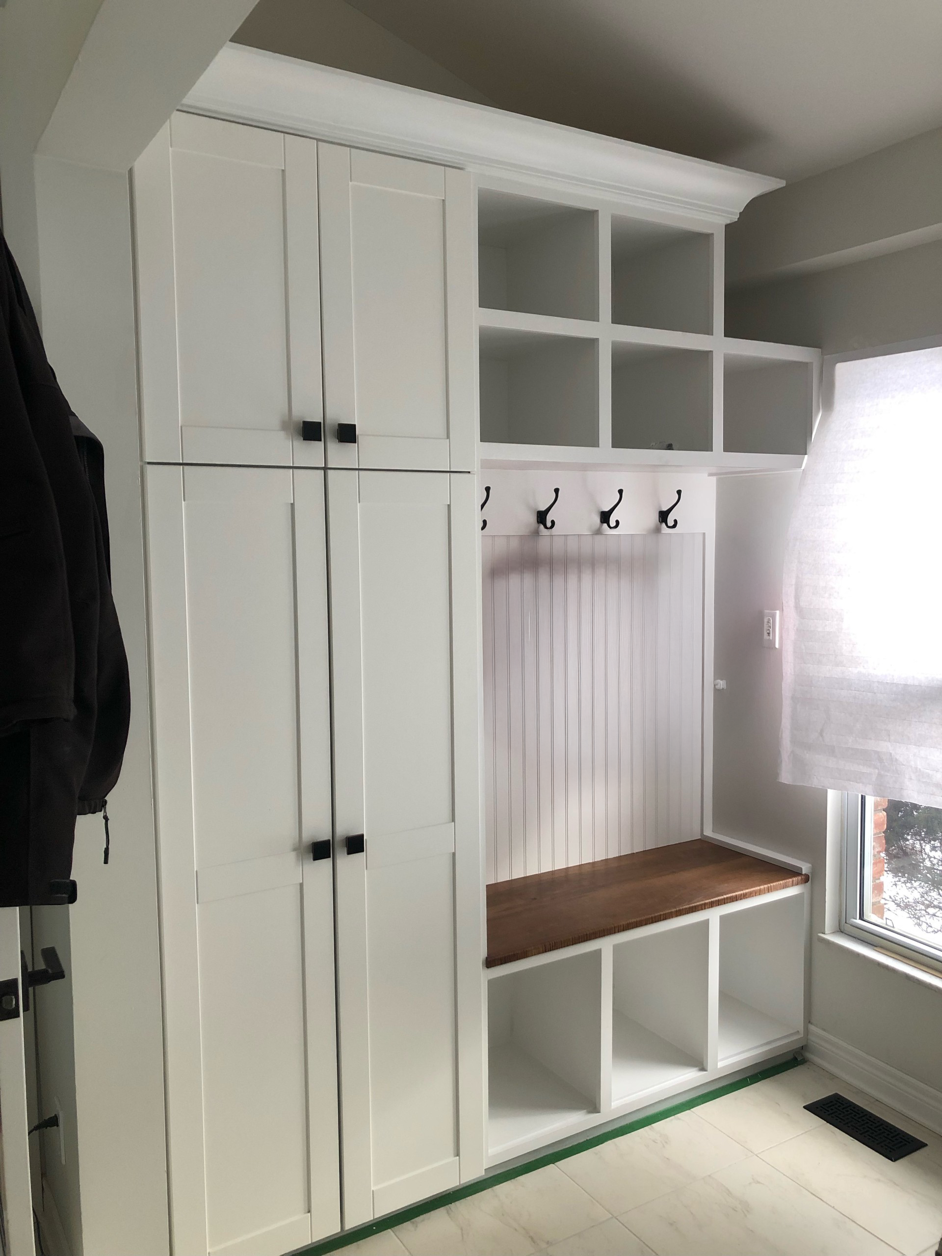 White built-in mudroom cabinet with cubbies, hooks, bench, and storage cupboards beside a window.