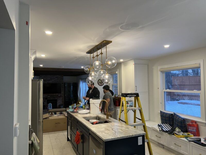 Two people working in a modern kitchen with an island, pendant lights, and a ladder near the window.