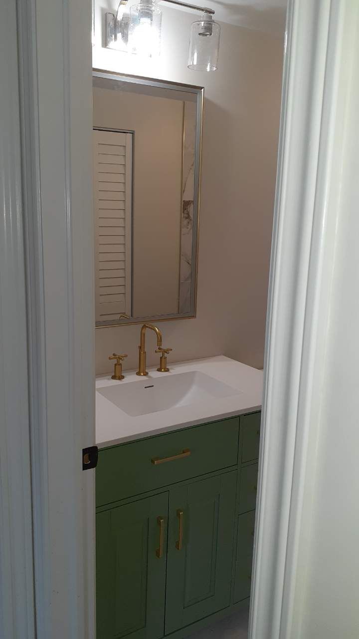 Small bathroom vanity with green cabinet, white sink, gold faucet, and mirror under a light fixture.