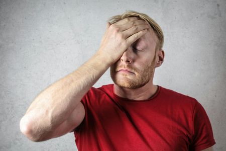 A person in a red t-shirt stands against a grey wall with their hand over their face in a gesture of frustration.