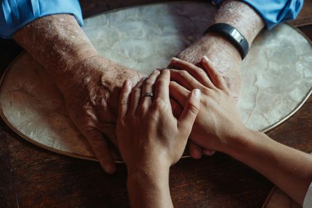 Two pairs of hands, one weathered and one smoother, resting together on a textured placemat over a dark table.