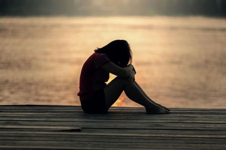 A person sitting with their head tucked into their knees on a wooden dock by calm water at sunset.