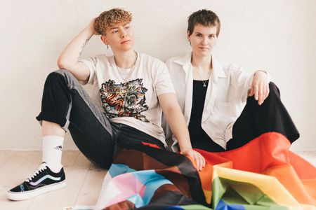 Two people sit against a white wall, draping a pride flag over their laps. They look toward the camera.