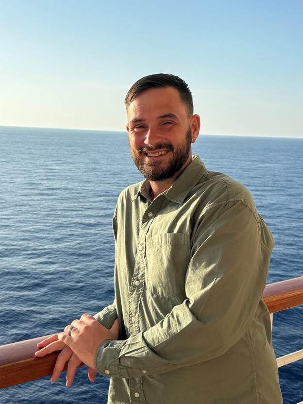 A person with a short beard smiles while standing at a wooden railing, overlooking the open sea under a clear sky.