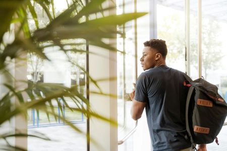 A person with a backpack stands inside a sunlit doorway, looking toward the exterior.
