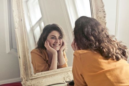 A person in a yellow top smiles at their reflection in an ornate, vintage-style floor mirror.