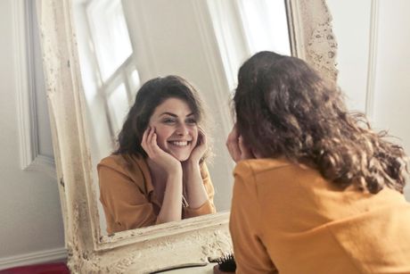A person in a yellow top smiles at their reflection in an ornate, vintage-style floor mirror.