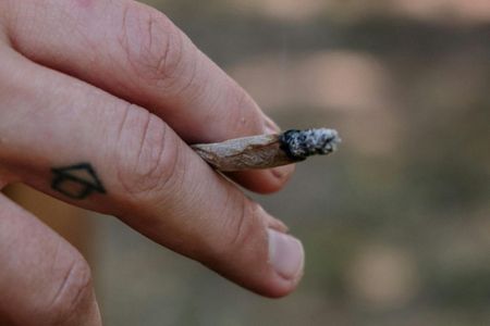 A close-up view of a hand holding a lit, hand-rolled cigarette, with a small house-shaped tattoo on a finger.