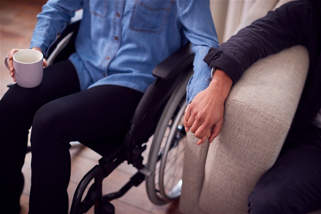 Woman pushing a person in a wheelchair on a sunny street, both smiling with outstretched arms.