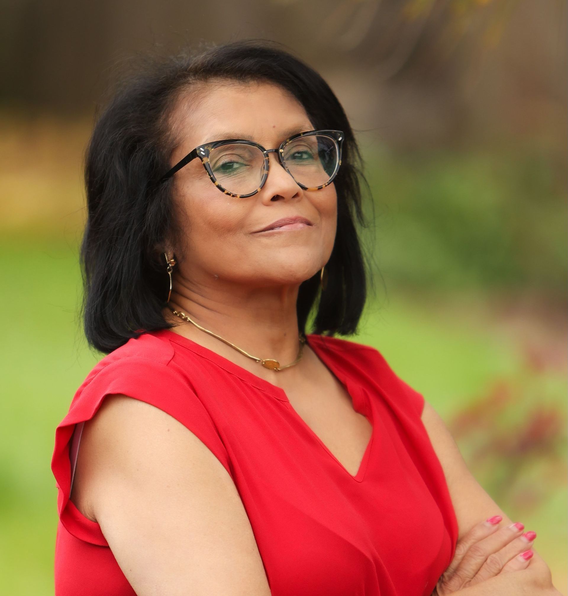 A woman wearing glasses and a red dress is standing with her arms crossed.