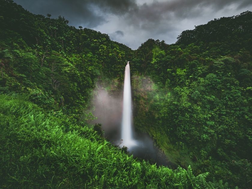 Waterfall cascading into a pool, surrounded by lush green vegetation under a cloudy sky. Private oceanfront stay Big Island