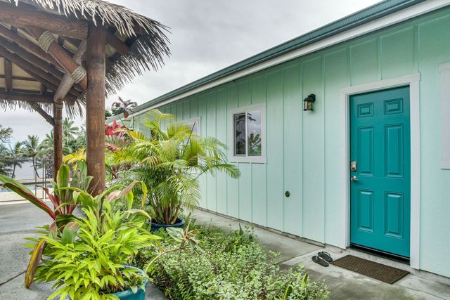 Teal door and window on a light blue building, next to a thatched roof structure and plants.