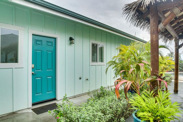 A turquoise door and windows on a light green building, with plants and a thatched-roof structure alongside.