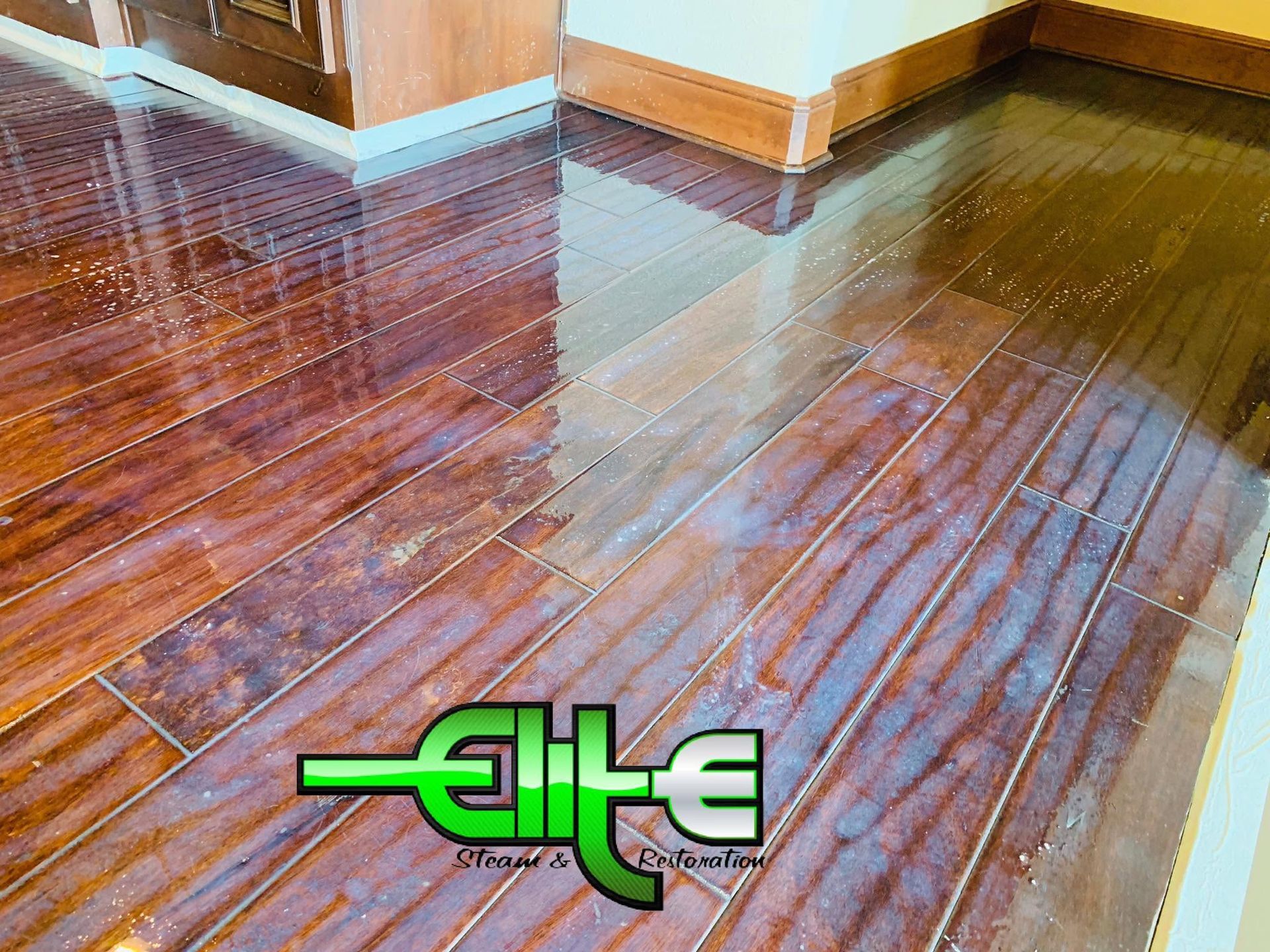Shiny wooden floor in a room corner, reflecting light, with green logo text near the bottom.