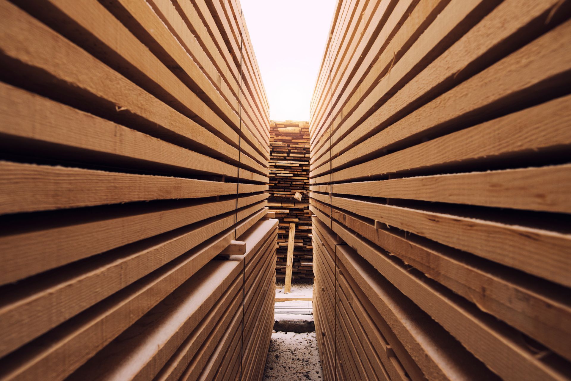 Stacks of lumber, light brown, filling the frame with a narrow pathway leading to the light in the distance.