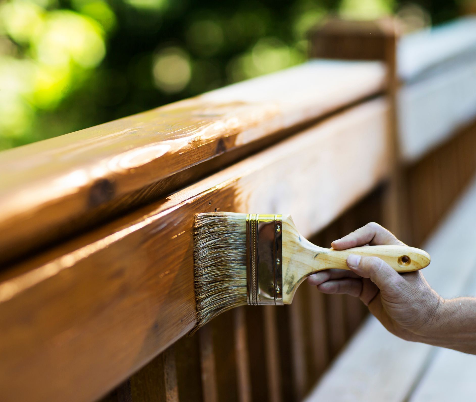 Hand painting a wooden deck railing with a brush and brown stain, outdoors.