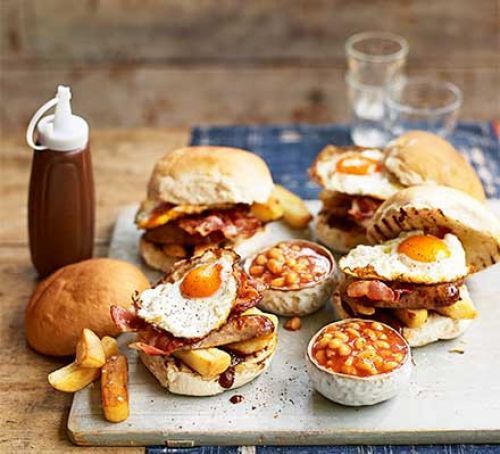A bunch of sandwiches with eggs , bacon , beans and potatoes on a table.