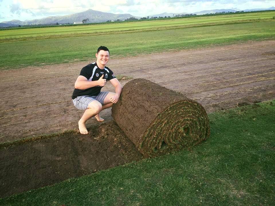 A Man Is Squatting On Top Of A Roll Of Turf In A Field — Marian Lawns In Marian, QLD