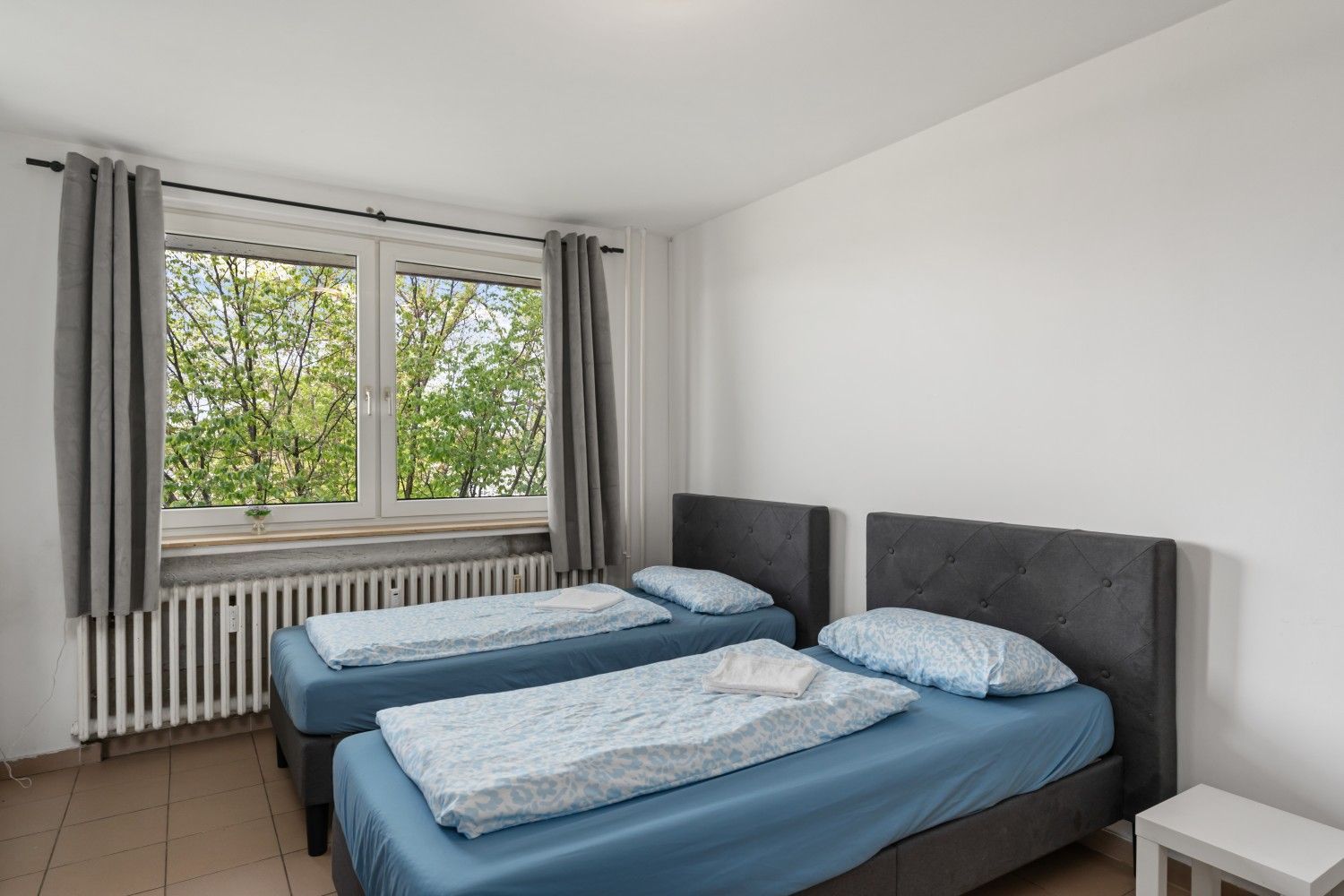 Helles Schlafzimmer mit zwei blauen Betten, grauen Vorhängen und einem großen Fenster mit Blick auf die Bäume
