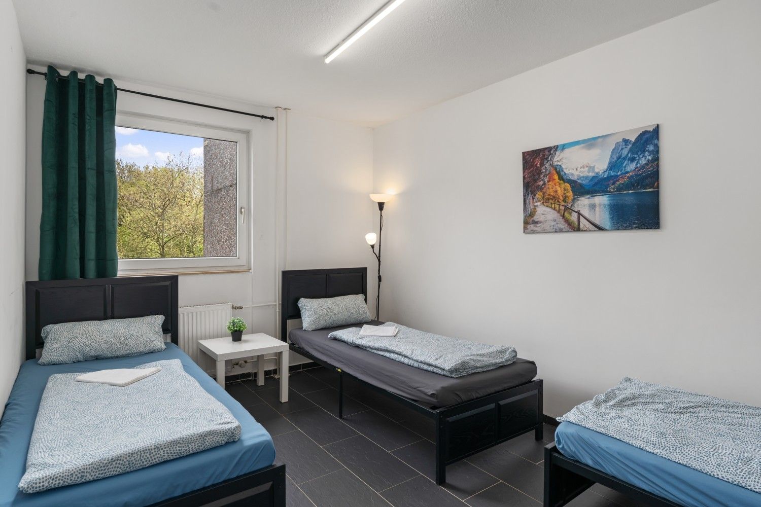 Kleines Schlafzimmer mit drei Einzelbetten, einem Fenster und einem an der Wand befestigten Landschaftsgemälde.