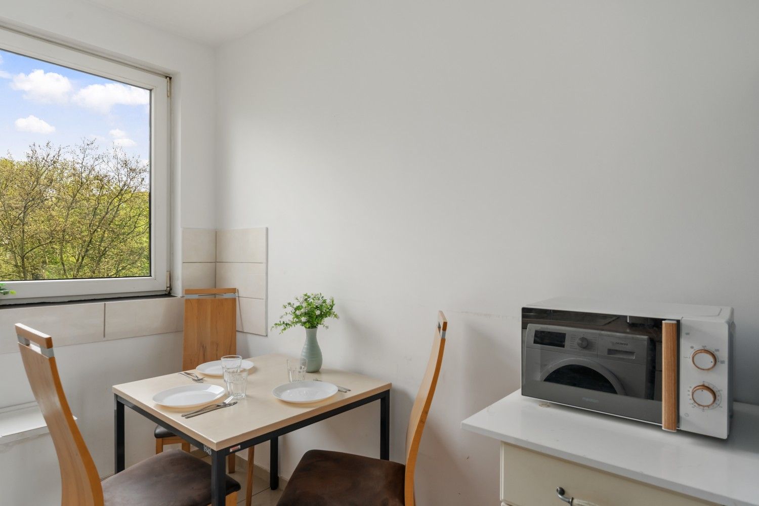 Kleine Essecke mit Tisch und Stühlen am Fenster und einer Mikrowelle auf einem Schrank.