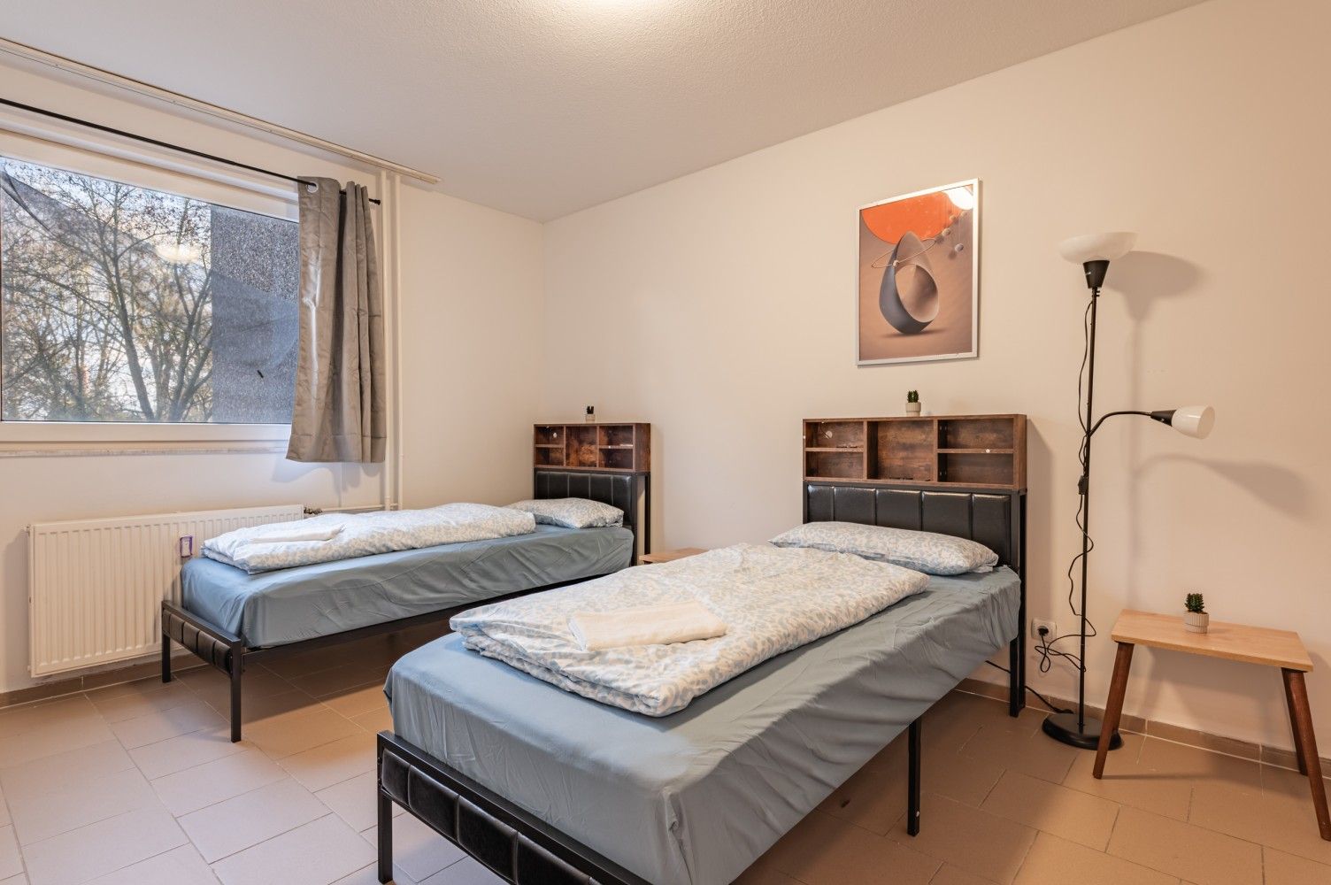 Schlafzimmer mit zwei Einzelbetten, Fenster, Stehlampe und kleinem Beistelltisch in einem hellen Zimmer