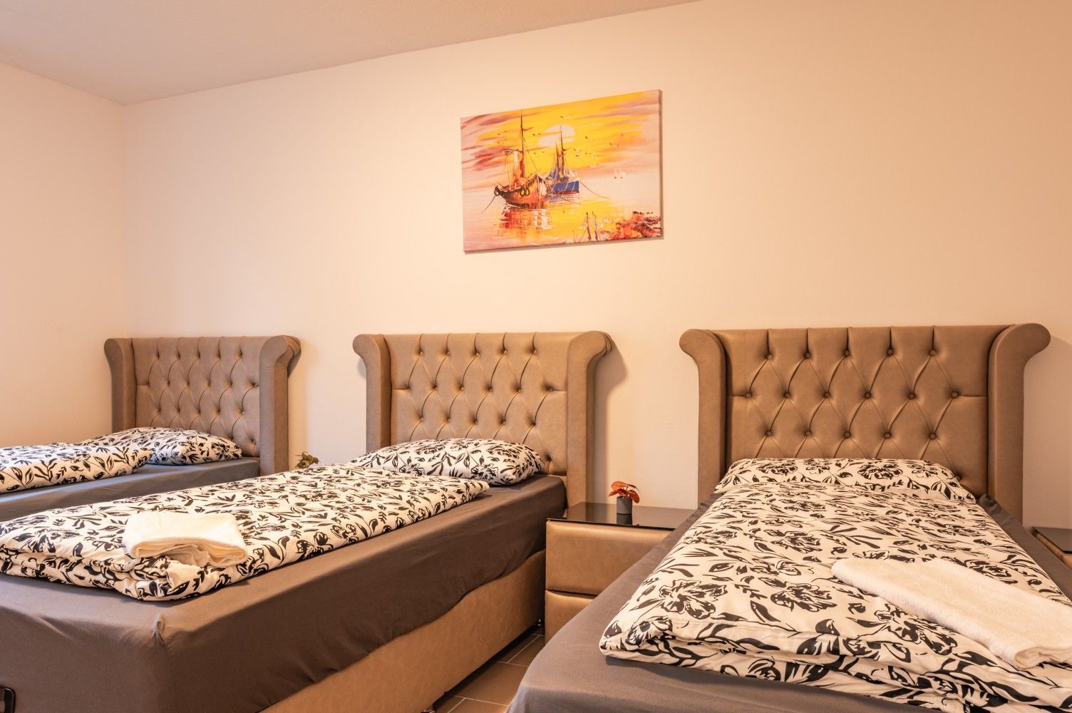 Schlafzimmer mit drei Einzelbetten, beigefarbenen, gesteppten Kopfteilen und einem kleinen Gemälde über dem mittleren Bett.