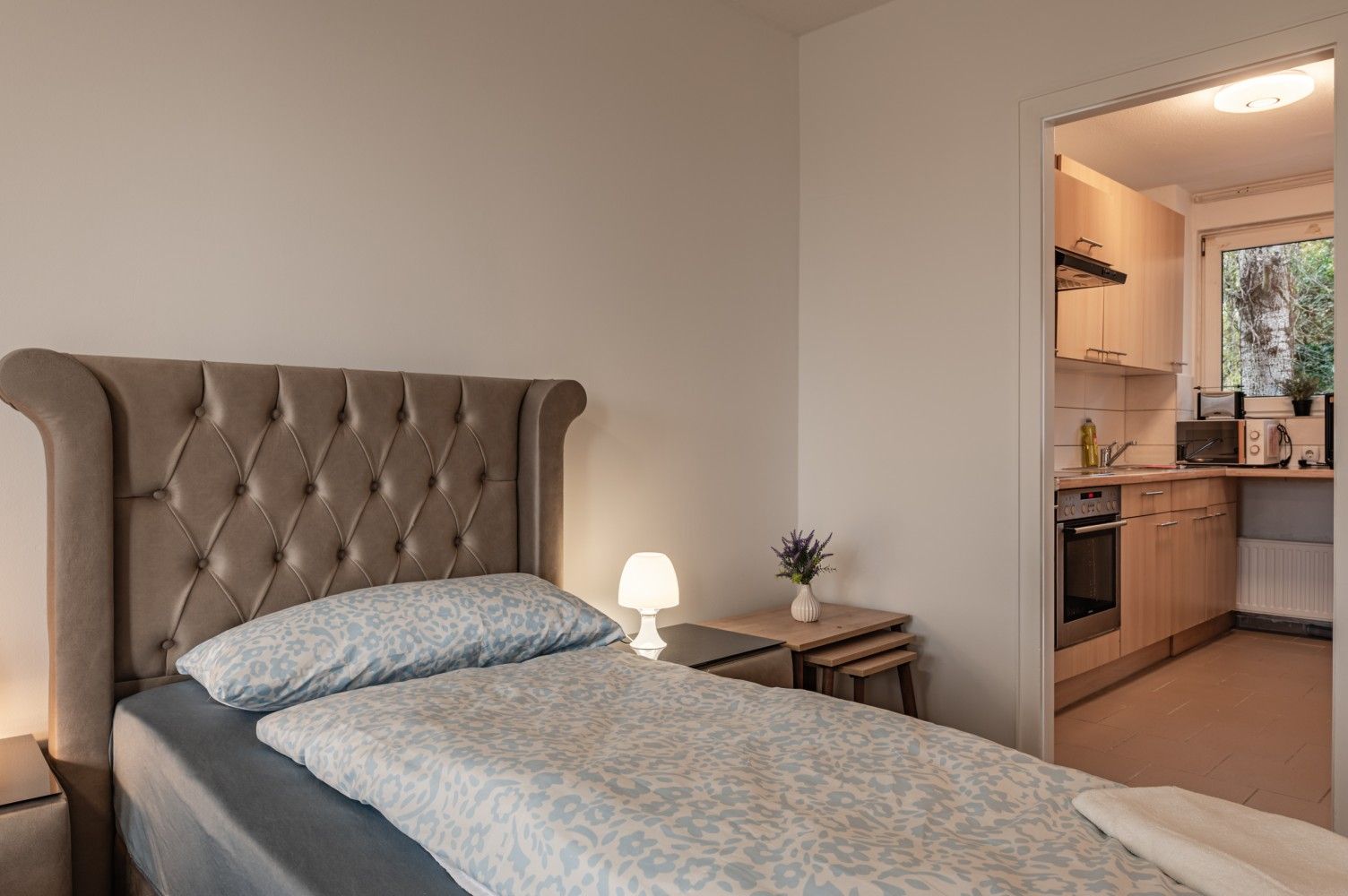 Gemütliches Schlafzimmer mit gestepptem grauen Kopfteil, Nachttischlampe und Blick in eine kleine Küche.