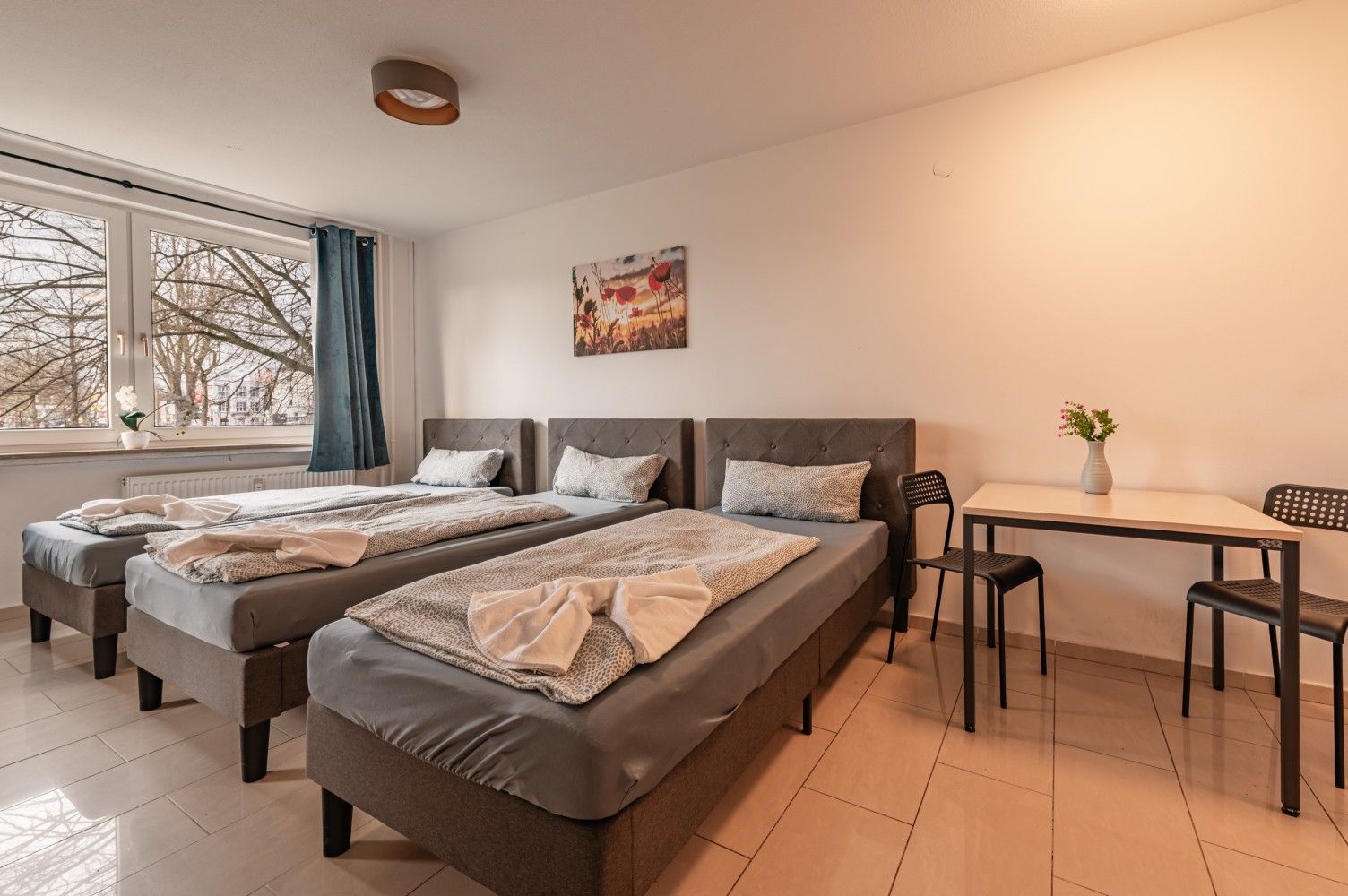 Helles Schlafzimmer mit zwei Betten, einem Fenster und einem kleinen Tisch mit Stühlen und Blumen.