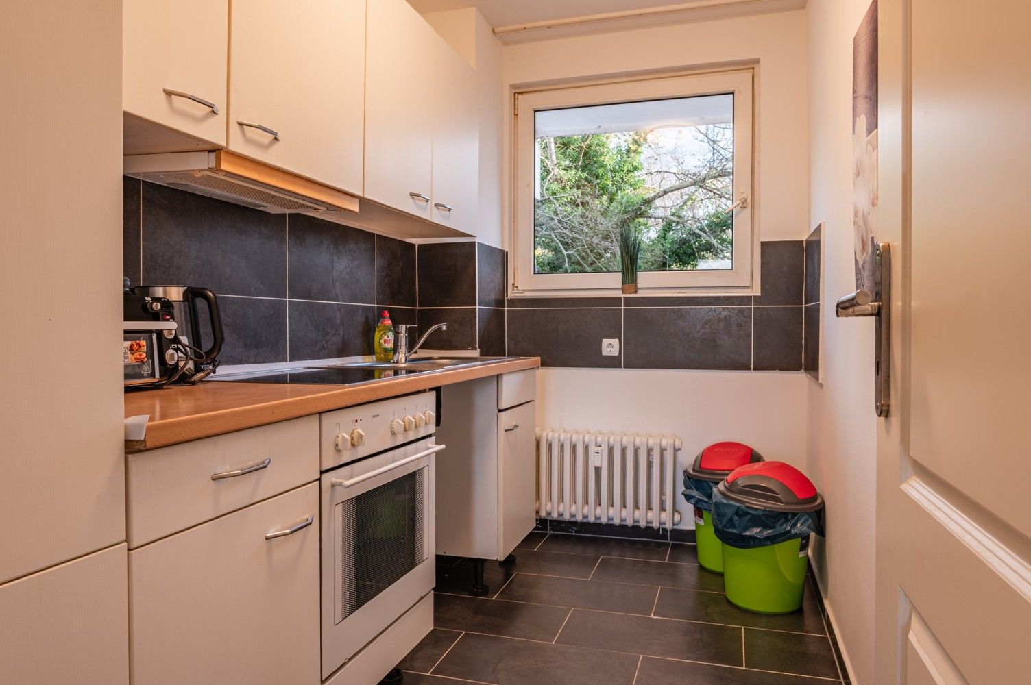 Schmale Küche mit weißen Schränken, schwarzem Fliesenspiegel, Fenster, Backofen, Spüle und Putzeimern auf dem Boden.