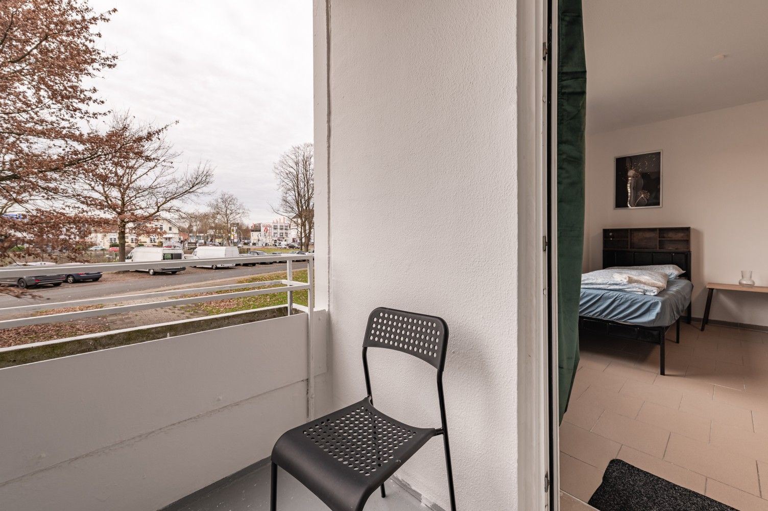 Blick vom Balkon durch eine offene Tür in ein Schlafzimmer mit Bett und Kommode; auf dem Balkon steht ein Stuhl.