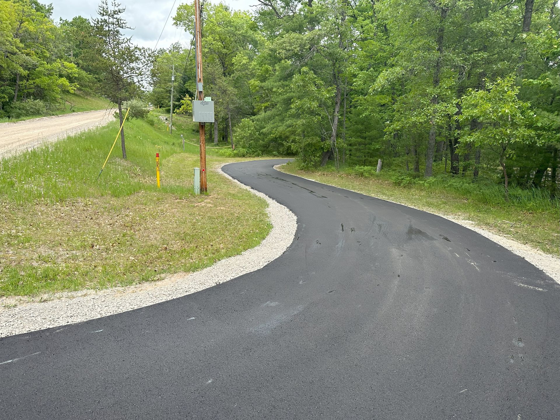 Curved freshly paved black road through green trees and grass, with a gravel shoulder and signpost