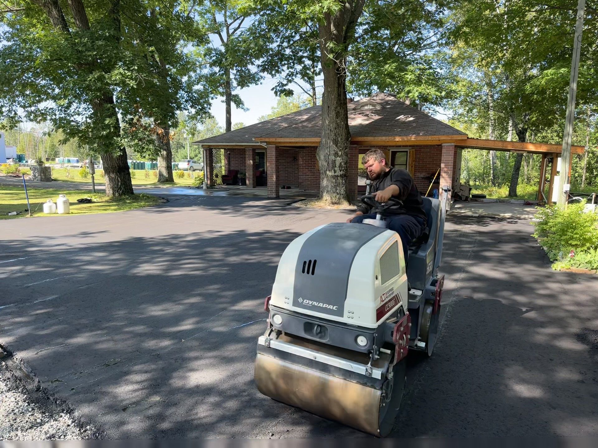 Person driving a ride-on street sweeper on a paved path beside a park building and trees
